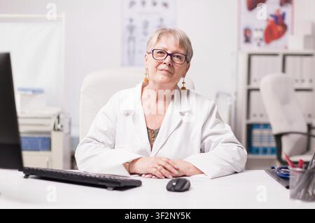 Confident Senior Female Doctor in Clinic wearing glasses, wearing a white coat, smiling at camera in a hospital cabinet, presenting a professional healthcare portrait in a clinical setting. Stock Photo