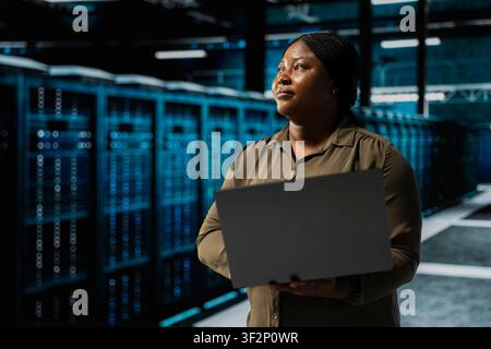 Programmer holding laptop walking through server room rows lined with storage rigs. IT expert using notebook program to inspect data center helping power internet services for business clients Stock Photo