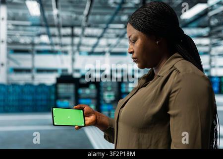 Woman in data center monitoring equipment parts for potential failures using green screen smartphone. African american server hub worker uses mockup phone to configure settings, increasing uptime Stock Photo