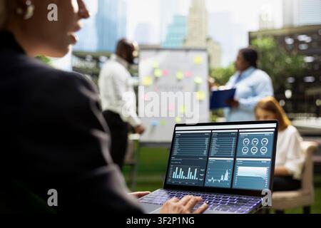 Businesswoman using laptop displaying financial dashboard in outdoor workshop, data analytics and metrics for business performance. Trading chart indicators on device screen. Stock Photo