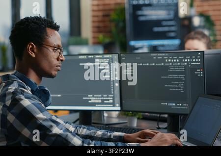 Software developer thinking while touching beard while typing on laptop sitting at desk with multiple screens parsing code, focused database admin working with team coding in the background. Stock Photo