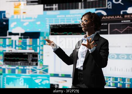 Manager hosting business meeting with shareholders, showing performance metrics graphs on screen. Businesswoman briefing management executives, holding speech, talking in mic Stock Photo