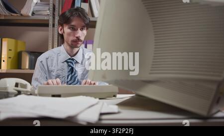 Office worker typing at cluttered desk with oversized monitor in compact records room, showing focused data entry amid papers, folders, binder Stock Photo