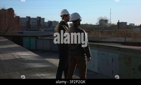 Two Engineers On Rooftop Review Plans On Tablet While Discussing Structural Details And Next Steps. Media Stock Photo