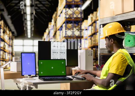 Male warehouse clerk printing barcode labels next to chroma key displays, handling tracking numbers labeling operations to prepare parcel shipment. Managing order fulfillment in depot. Stock Photo
