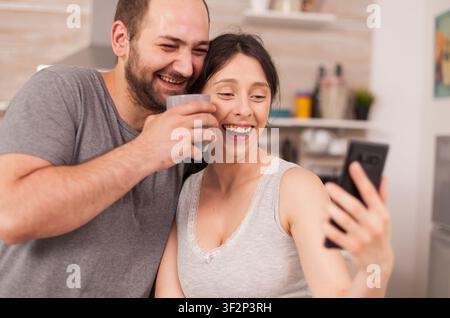 Happy young couple takes selfies during breakfast, making funny faces together in the kitchen and enjoying a joyful married morning. Stock Photo