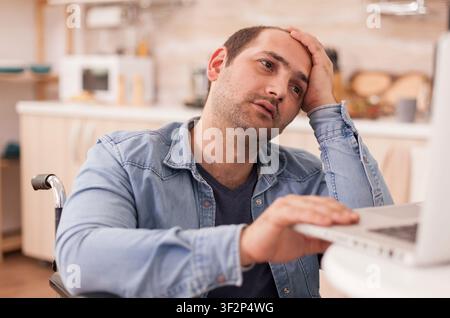 Stressed businessman with walking disability in wheelchair using laptop in kitchen, while disabled paralyzed handicapped man with walking disability integrating after an accident. Stock Photo