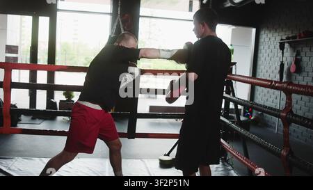 Two men sparring inside boxing ring as one boxer drives straight punches, retreats under pressure, while trainer holds focus mitts in bright backlit Stock Photo