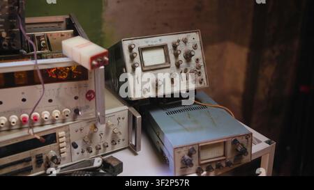 Vintage analog oscilloscopes and test instruments stacked on cluttered workshop bench under warm directional light, with worn cables, knobs, metal Stock Photo