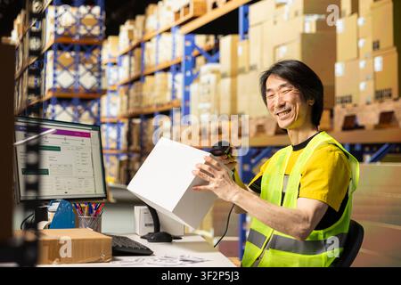 Joyful asian man using a scanner to register orders for tracking, handling cargo goods preparations before shipment in a distribution hub. Packaging, tagging and scanning products. Stock Photo