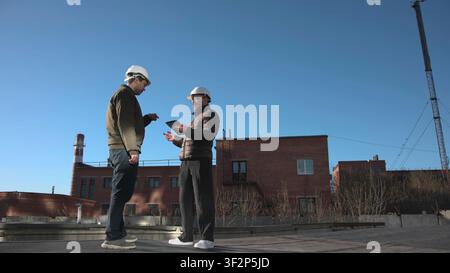 Group Of Construction Laborers Carefully Analyzing Blueprints On Rooftop Amidst Clear Sky Conditions. Media Stock Photo