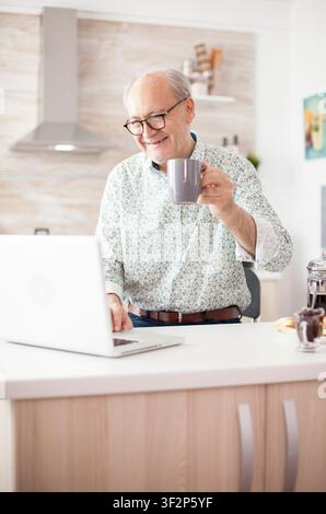 Cheerful senior man enjoys breakfast and coffee while greeting family through a laptop video call in the kitchen, during a breakfast routine in a home kitchen. Stock Photo