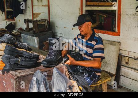 Balikpapan, Indonesia - June 3 2026. An elderly Indonesian shoemaker wearing a cap and striped t-shirt, focused on fixing a heavy black combat boot. Stock Photo