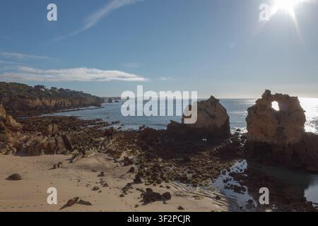 The scene shows rock formations on a beach with waves lapping at the shore. The sun shines brightly overhead while clouds drift in the distance. Stock Photo