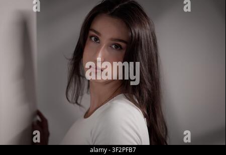 Young woman posing against a light wall, looking at the camera with a calm expression Stock Photo