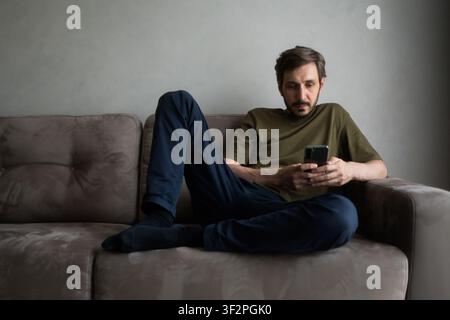 Man relaxing on a living room sofa, feeling isolated and sad while engrossed in his mobile phone, reflecting concepts of technology addiction, depress Stock Photo