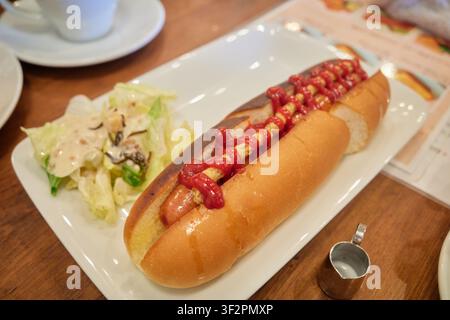 A grilled sausage hot dog with ketchup and a side salad on a wooden cafe table perfect for regional food blogs and menu marketing. Stock Photo