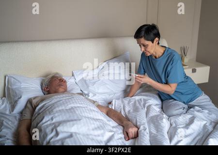 Concerned senior woman checking temperature level with thermometer of sick partner lying in bed Stock Photo
