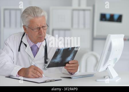 doctor examining x-ray image and taking notes in medical office with computer monitor and stethoscope around neck, professional setting, white and Stock Photo