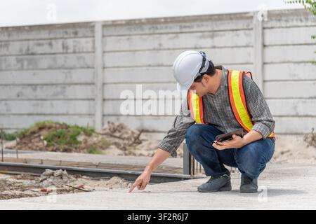 Engineer inspecting concrete surface at construction site while using tablet and safety equipment for quality control structural assessment and projec Stock Photo