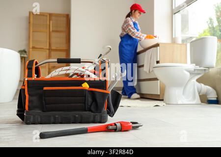 Plumbing toolkit with wrench and pipe wrench on bathroom floor while female plumber works on sink, a female plumber in blue overalls and a red cap wor Stock Photo