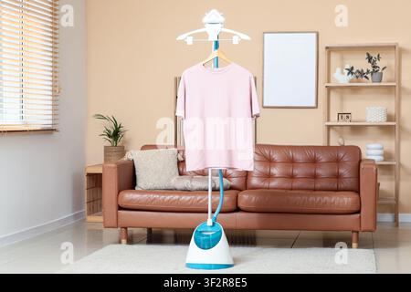 Pink t-shirt on a clothes steamer in a modern living room with a brown leather sofa and wooden blinds Stock Photo