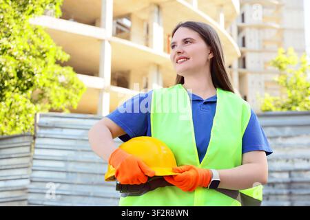 Smiling female construction worker holding a yellow hard hat and wearing safety gloves at a building site, the woman looks confidently to the side, pr Stock Photo