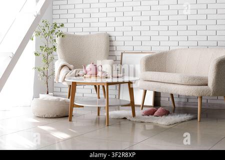 Cozy living room interior with a comfortable armchair, sofa, coffee table, and houseplant against a white brick wall Stock Photo