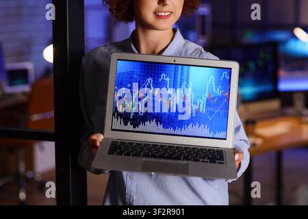 Smiling woman holding a laptop displaying financial stock market charts and graphs in a dark office environment, the scene takes place in a dark offic Stock Photo