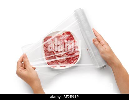 Hands wrapping a plate of sliced sausage and ham with plastic cling film for food preservation isolated on white background Stock Photo