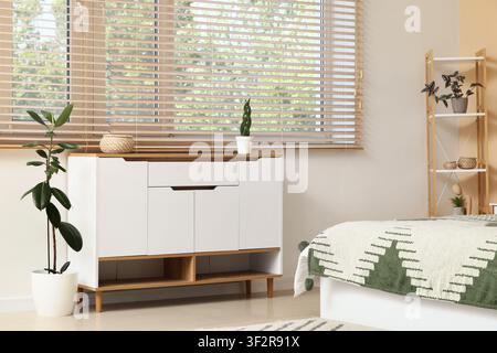 Modern bedroom interior with white and wood dresser, green plant, and bed with patterned blanket Stock Photo