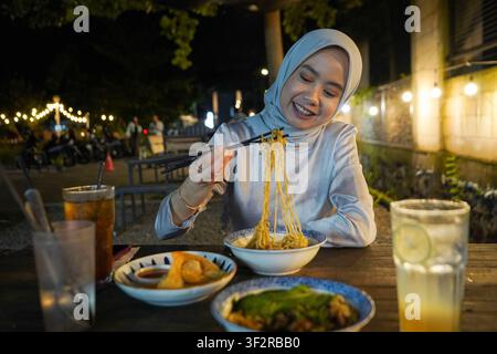 Joyful Asian Muslim woman in hijab eating noodles with chopsticks at an outdoor night market food stall Stock Photo