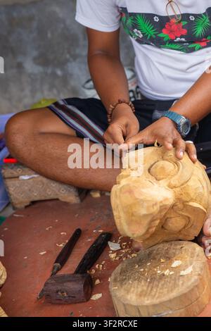 A skilled Balinese craftsman using a wooden mallet and chisel to shape a traditional mask from raw timber. Stock Photo