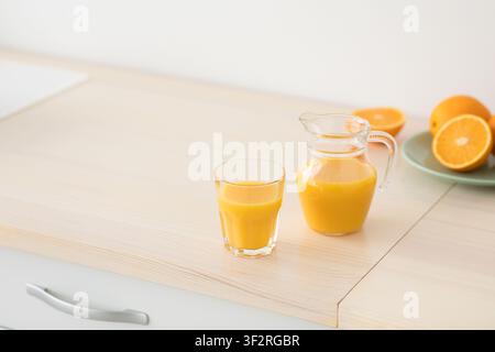 Freshly squeezed orange juice in glass and jug, fruits on plate on white furniture in modern minimalist kitchen Stock Photo