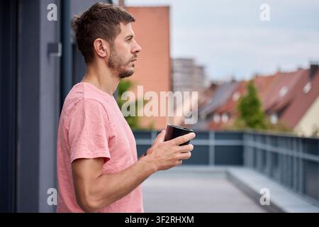 Man holding a coffee cup while standing on an apartment terrace in the morning. Man taking a break outdoors Stock Photo