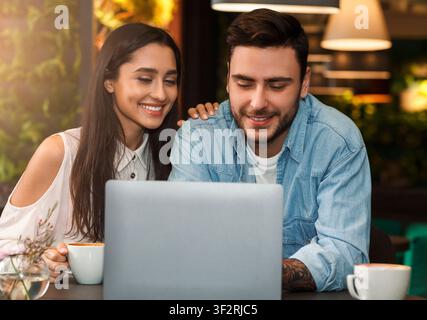 Cheerful Couple Using Laptop Having Coffee Sitting In Cafe Stock Photo
