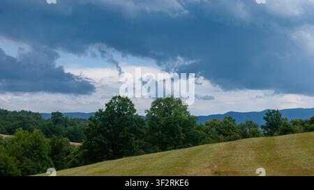 Dark storm clouds gathering above rolling countryside with green trees and open meadow in summer before rainfall. Stock Photo