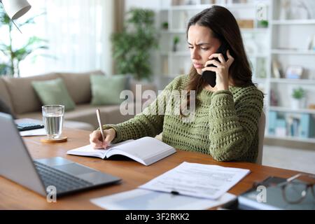 Annoyed teleworker talking on phone and checking agenda working at home Stock Photo