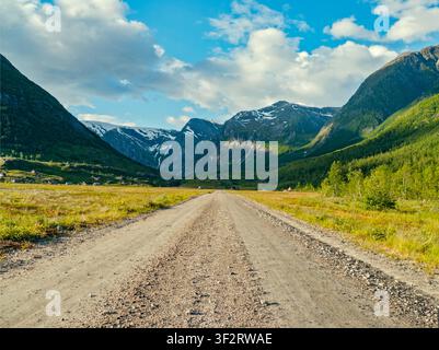 Veitastrond, Norway - 10.07.2017: A long dirt path cuts through verdant mountain meadows toward massive snow-dusted peaks under bright clouds Stock Photo