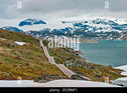 Luster, Norway - 12.07.2017: A winding road snakes through rugged mountain terrain with melting snow and a large lake under cloudy skies Stock Photo