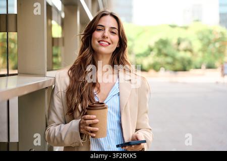 Smiling young businesswoman taking a break, holding a reusable coffee cup and smartphone, standing outside an office building Stock Photo