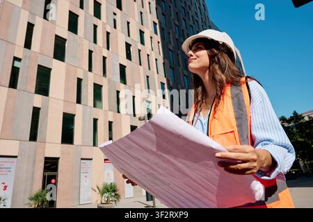 Young woman engineer wearing safety helmet and vest, reviewing construction plans outdoors on a sunny day Stock Photo