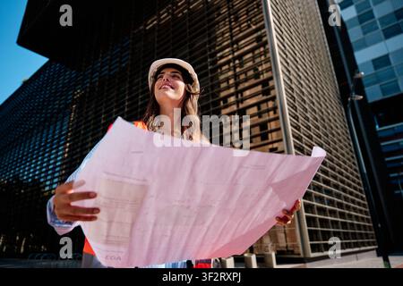 Female engineer wearing hard hat and safety vest, holding blueprints and looking up, imagining new building construction Stock Photo