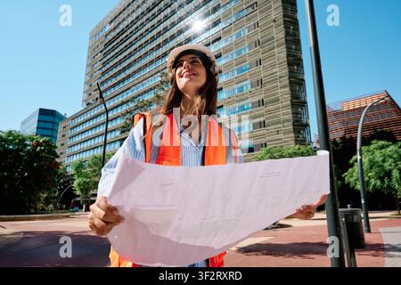 Young female engineer in hard hat and safety vest smiling, holding construction blueprints, standing in urban setting Stock Photo