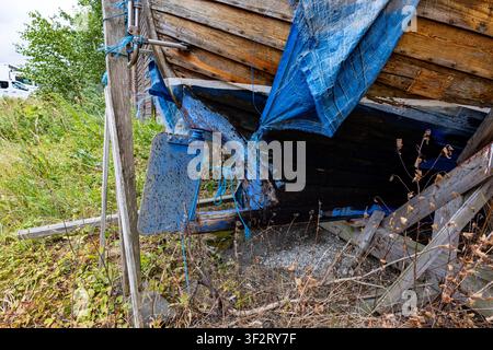 Blue tarp hangs torn from weathered wooden boat. Grass and fallen leaves surround the broken structure. Wooden planks show age and neglect. Background Stock Photo