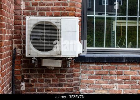 Outdoor air conditioner mounted on brick wall. Metal grille shields internal fan blades. Black-framed window beside unit. Weathered red bricks show ag Stock Photo