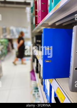 Blue binder stands out on a shelf filled with colorful folders in a stationery store, with a shopper walking in the background Stock Photo