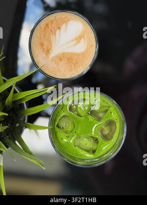 Top view of hot coffee with milk foam and bright green iced matcha drink on reflective cafe table. Modern beverage composition with plant leaves and Stock Photo