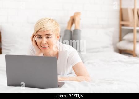Happy mature woman laying in bed and watching movie Stock Photo