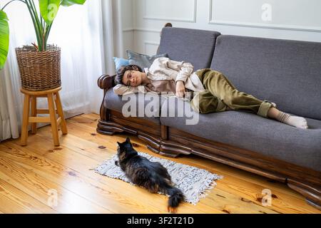 Lazy happy woman peacefully relaxing on couch and observing domestic cat resting on rug in room. Stock Photo